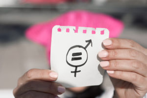 closeup of a young woman outdoors wearing a pink pussycat hat showing a piece of paper in front of her face with a symbol for gender equality drawn in it