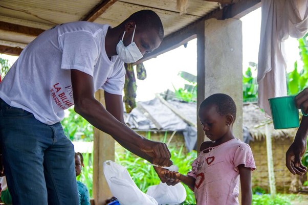 A man with a mask covering his moth and nose, is placing a medicine tablet into the hands of a young girl woth a pink shirt on. 