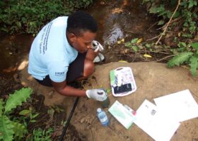 A woman wearing a light blue polo shirt with Citizen Researcher written on the sleeve, and white gloves, holding forceps over a container of pond water. She is squating down facing to the right and looking at a tray on the ground that has snall aquatic snails in it.