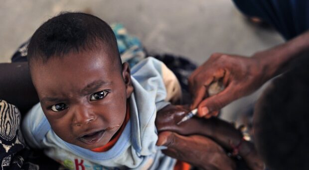 A young child, looking at the camera with a pained expression, being administered a vaccine in their arm.