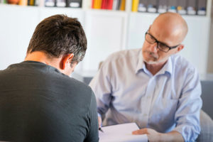 Mature male psychotherapist or counsellor interacts with a male client during a one-to-one therapy session.