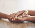 Close up of a child’s hand being held between the two hands of another person on a wooden table